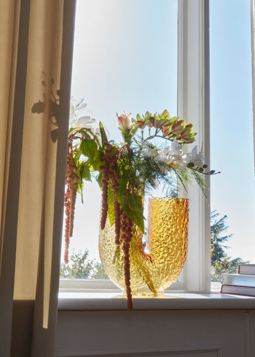 Wazon Arura High Amber wypełniony kwiatami na tle jasnych zasłon.